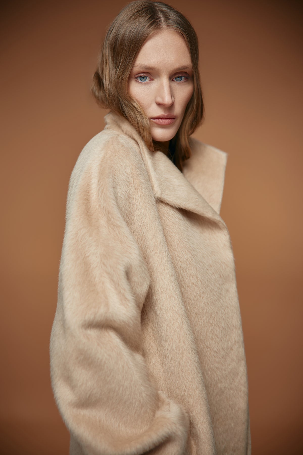 A woman wearing a plush, oversized beige faux fur coat, photographed against a solid warm brown studio background.