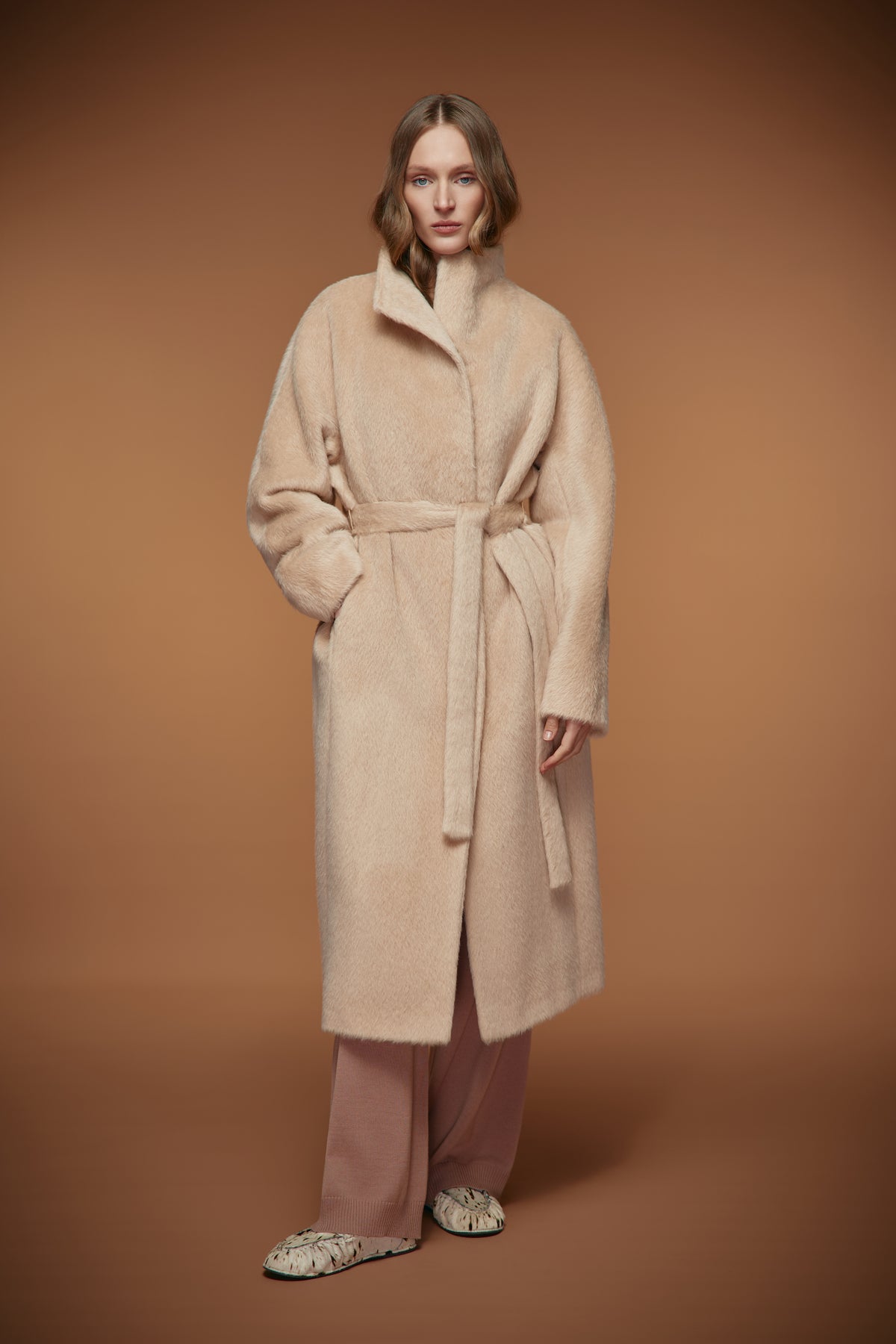 A woman stands against a warm brown studio background wearing a long cream-beige wool coat with a matching waist tie, dusty pink wide-leg trousers, and patterned slippers.