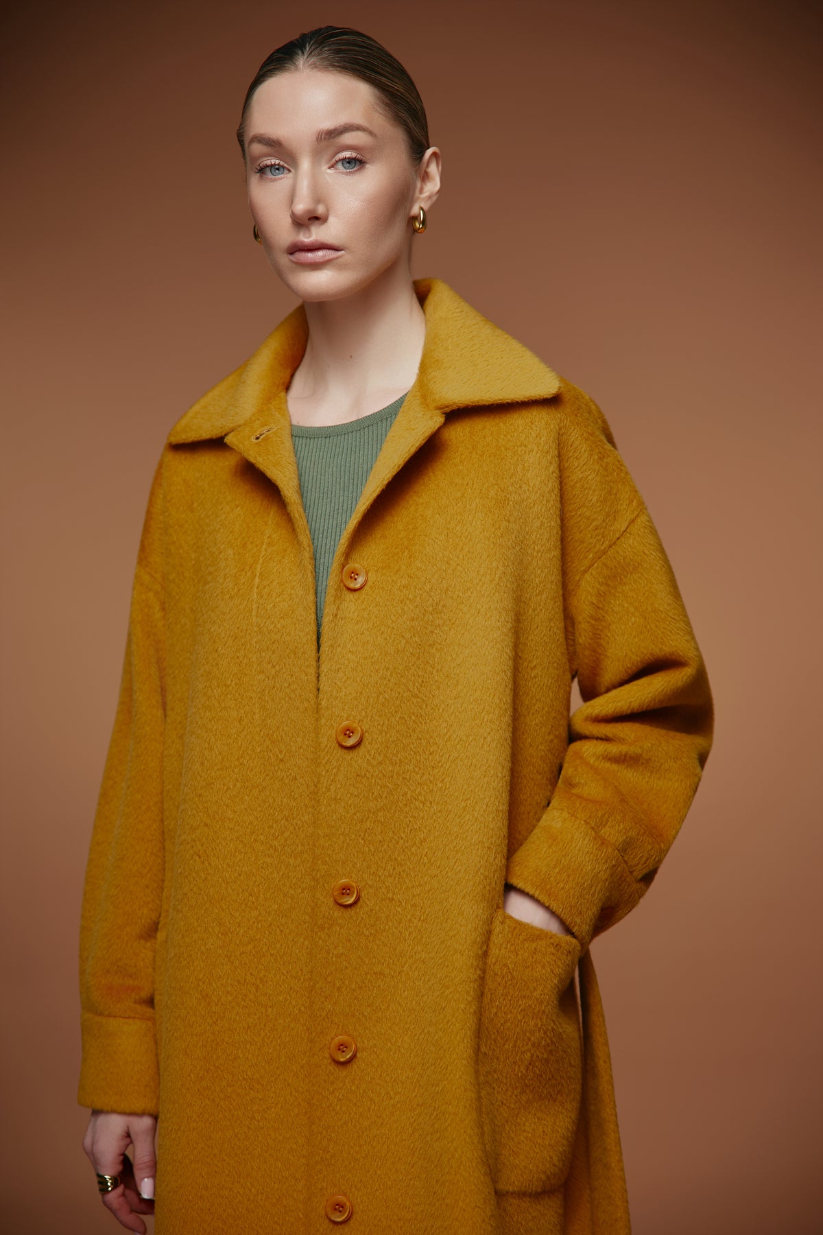 A woman modeling a long, oversized mustard yellow coat with a wide collar and front buttons, worn over an olive green top, set against a solid warm brown studio background.