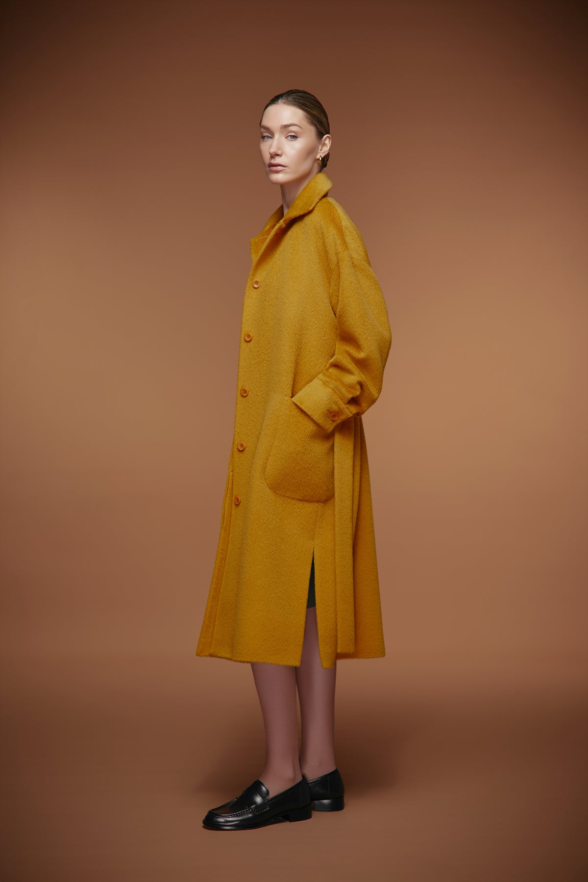 A model stands in profile wearing a long mustard yellow wool-blend coat with front buttons and large patch pockets, paired with black loafers, against a solid warm brown studio background.