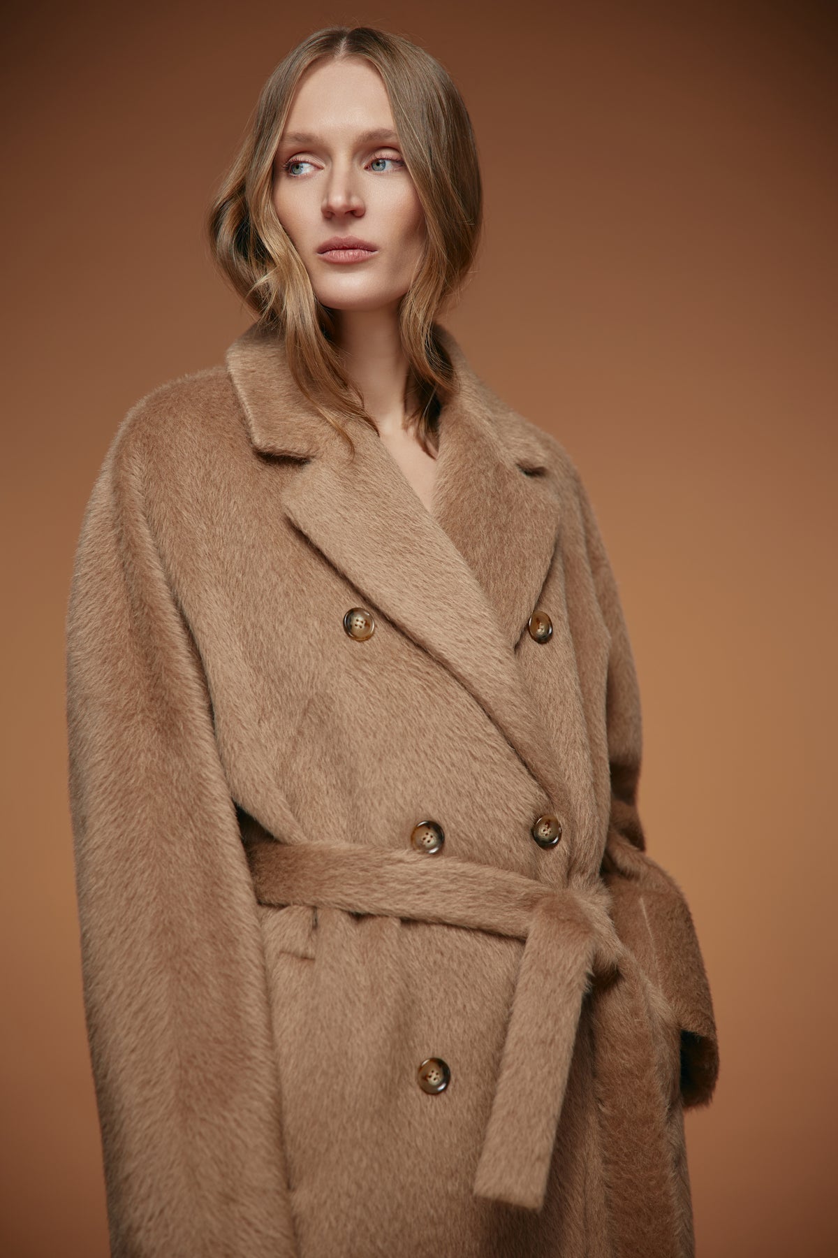 A woman wearing a plush camel-brown double-breasted faux fur coat with a matching waist tie, set against a solid warm brown studio background.