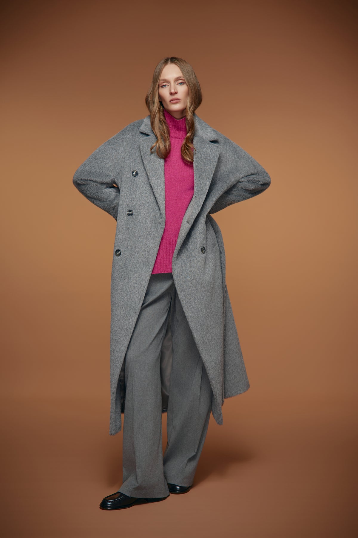 A woman modeling a long double-breasted gray wool coat over a bright pink turtleneck sweater, gray wide-leg trousers, and black loafers, set against a solid warm brown studio background.