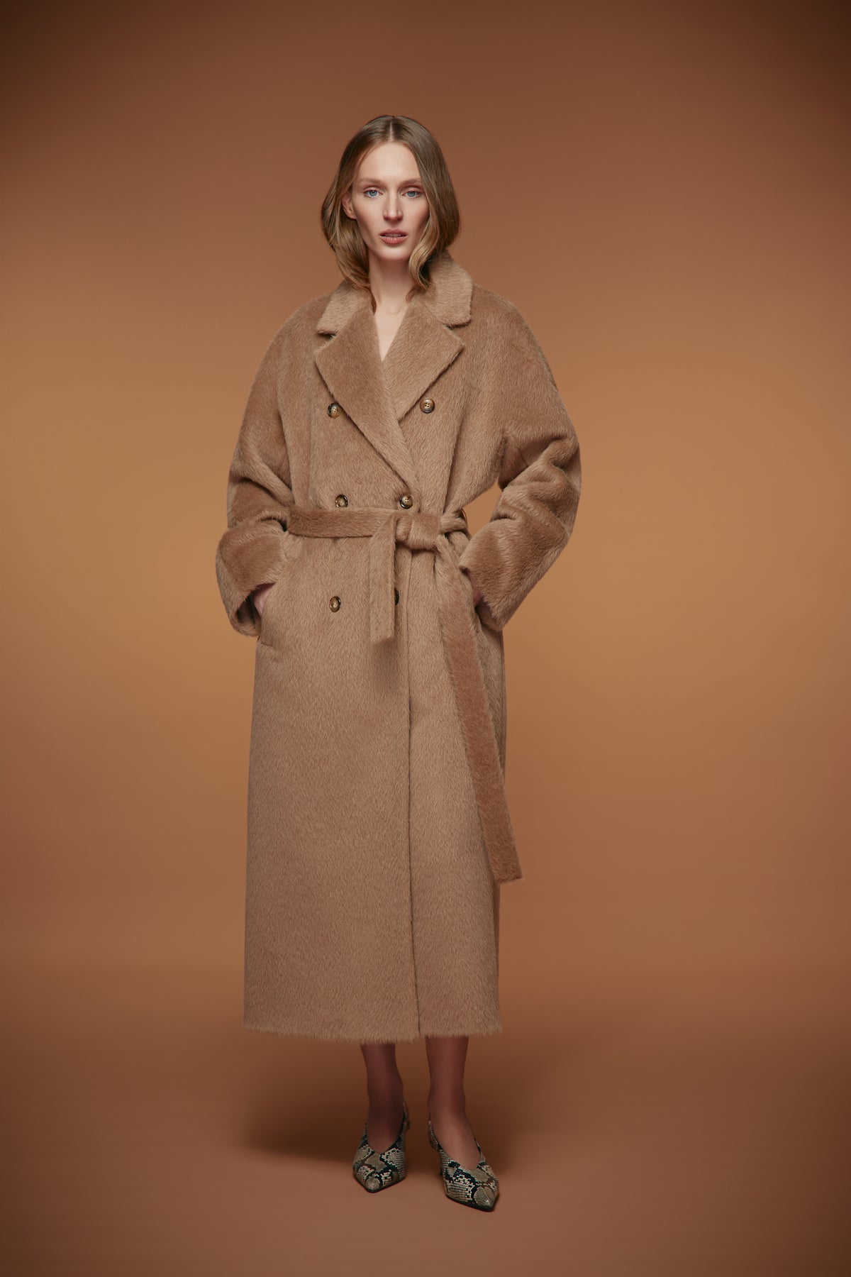 A woman modeling a long camel-brown double-breasted coat with a matching waist belt, standing against a solid warm brown studio background.