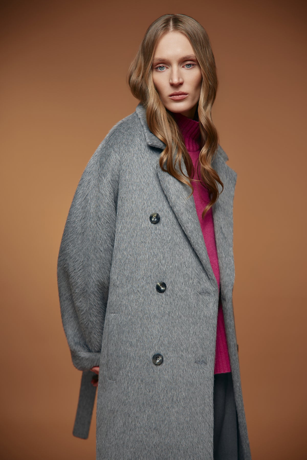 A woman modeling a long, double-breasted gray wool-blend coat with a belted waist, worn over a magenta turtleneck sweater, set against a solid warm brown studio background.