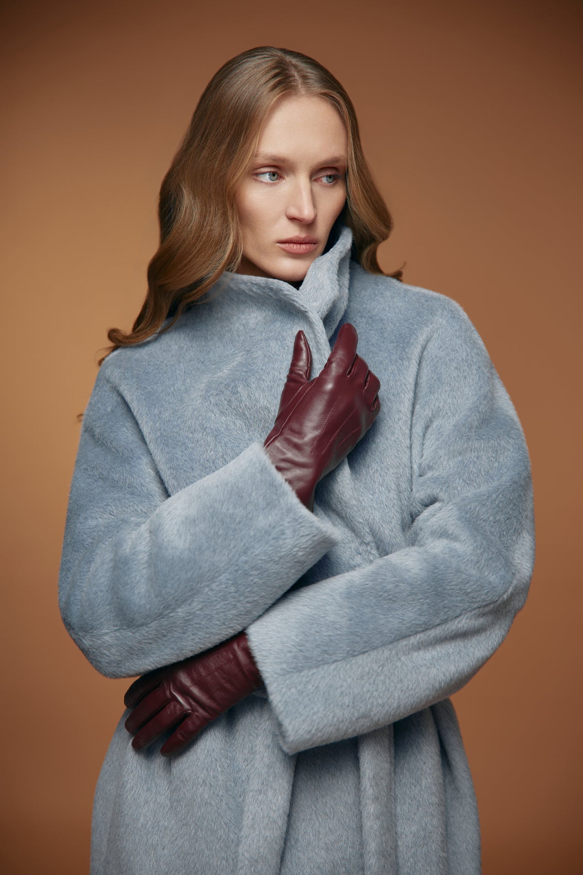 A woman wearing a light blue-gray fuzzy coat and dark burgundy leather gloves, posed against a solid warm brown studio background.