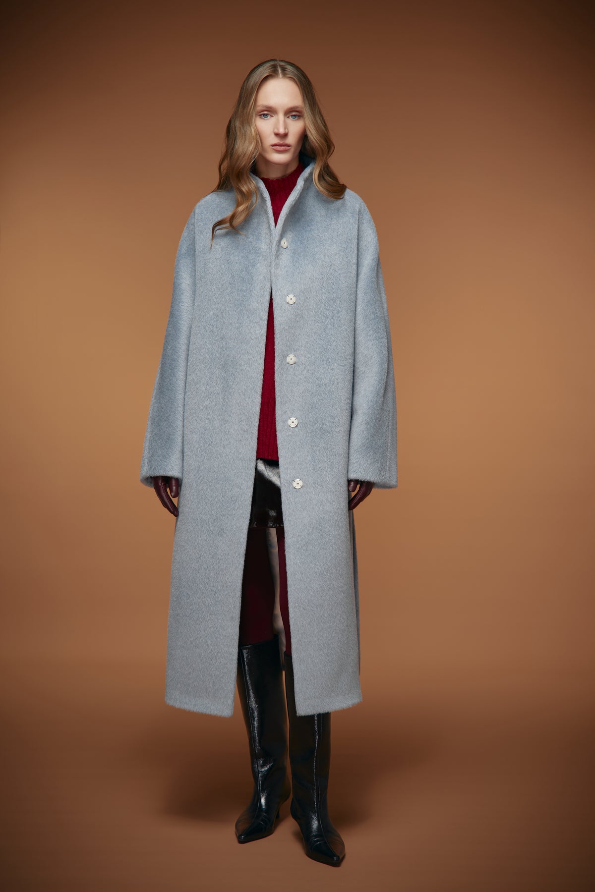 A woman stands against a warm brown studio background wearing a long light gray wool-blend coat over a burgundy turtleneck, dark pants, and black knee-high boots.