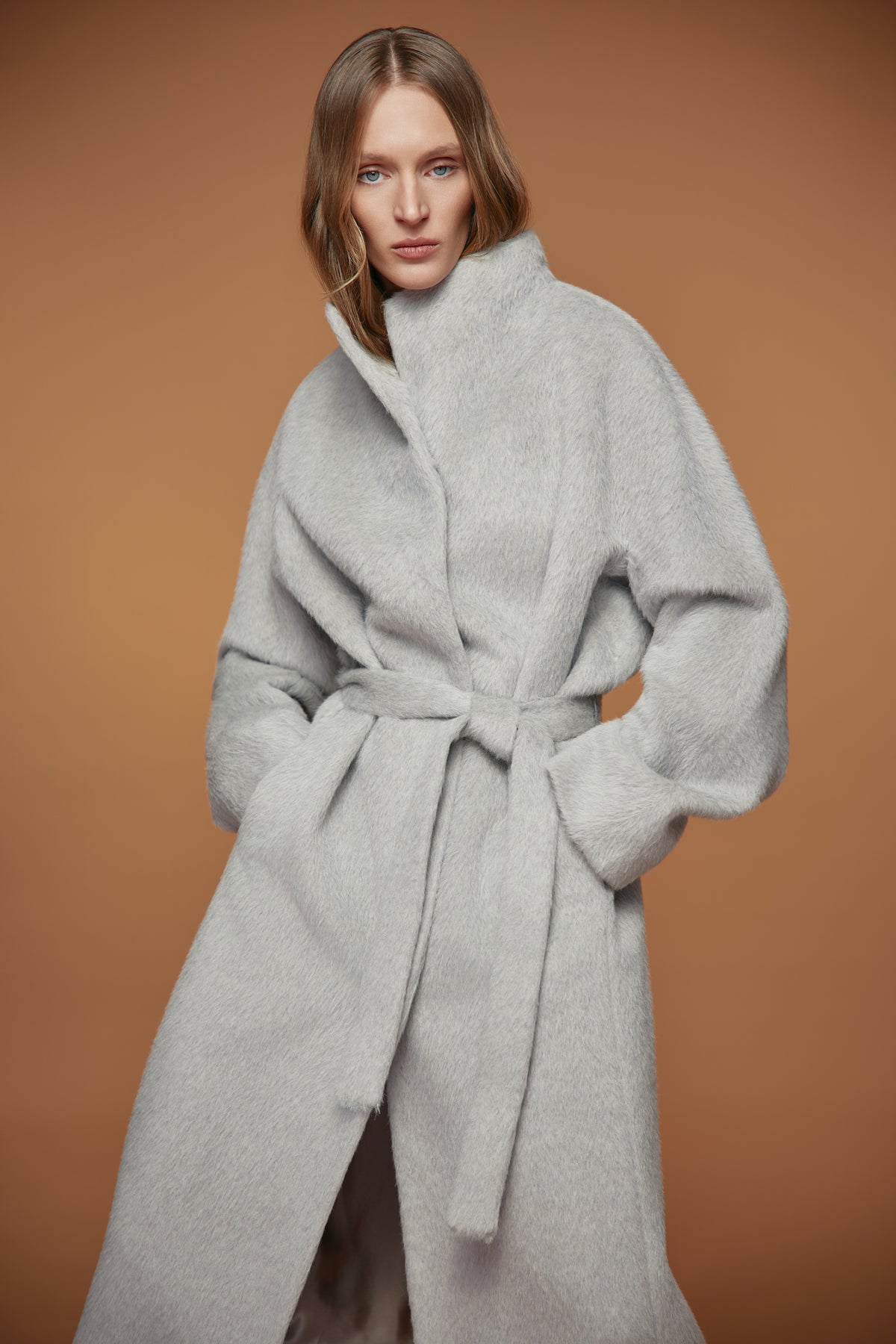 A woman wearing a light gray fuzzy belted coat against a warm brown studio background.