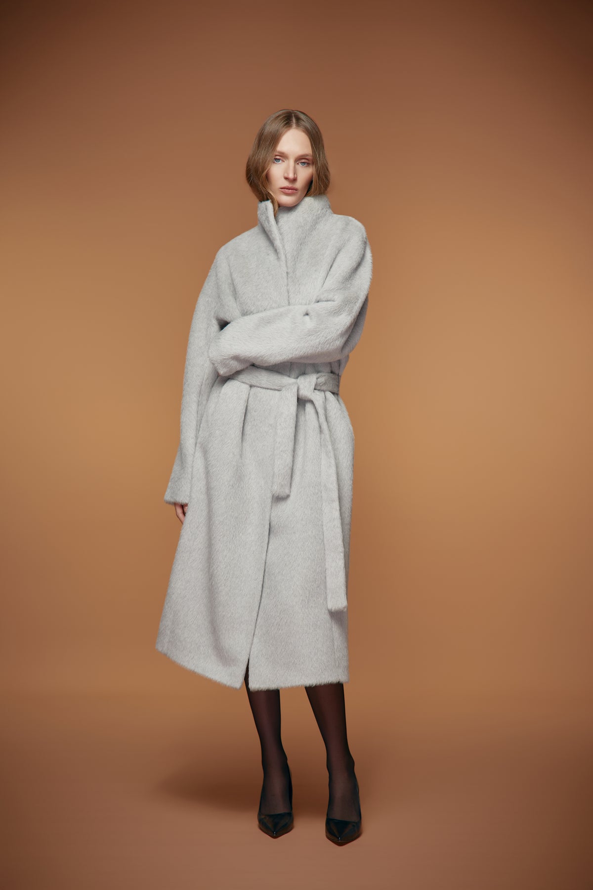 A woman modeling a light gray wool coat with a high collar and self-tie belt, paired with black tights and black pointed-toe heels, set against a solid warm brown studio background.