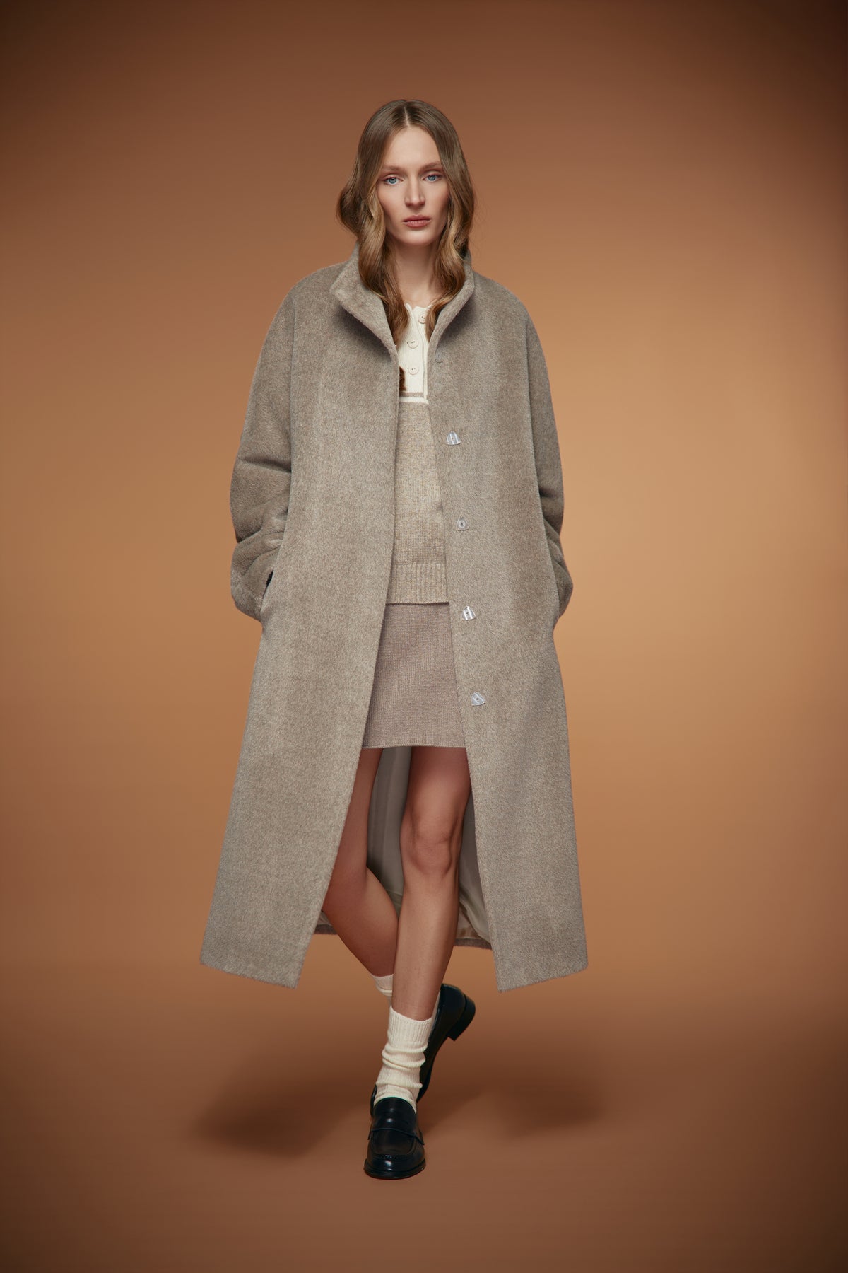 A model stands against a solid warm brown studio background wearing a long, oversized light beige wool-blend coat over a matching beige knit top and skirt, styled with cream socks and black loafers.