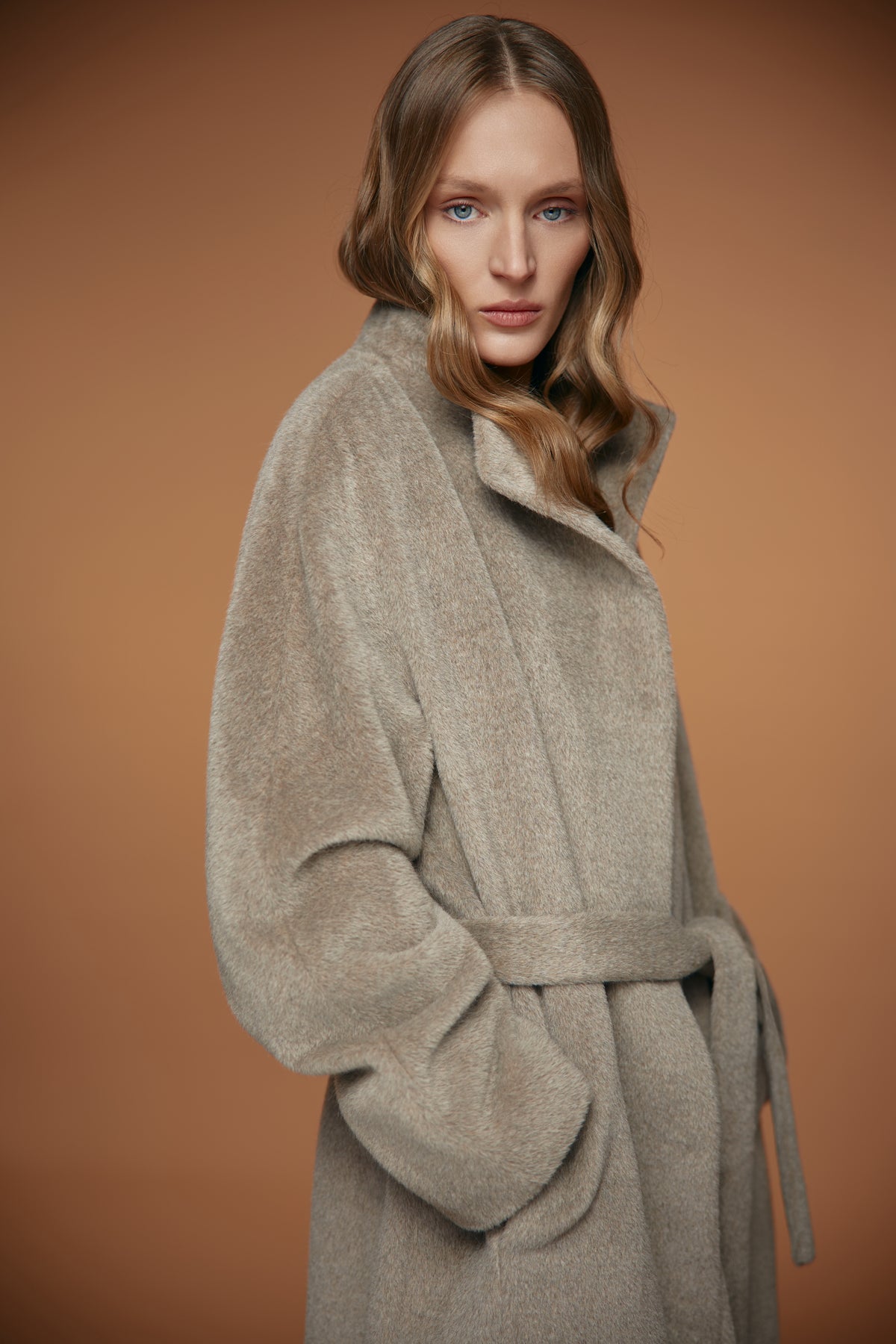A woman wearing an oversized, soft-textured beige coat with a matching belt, posed against a solid warm brown studio background.