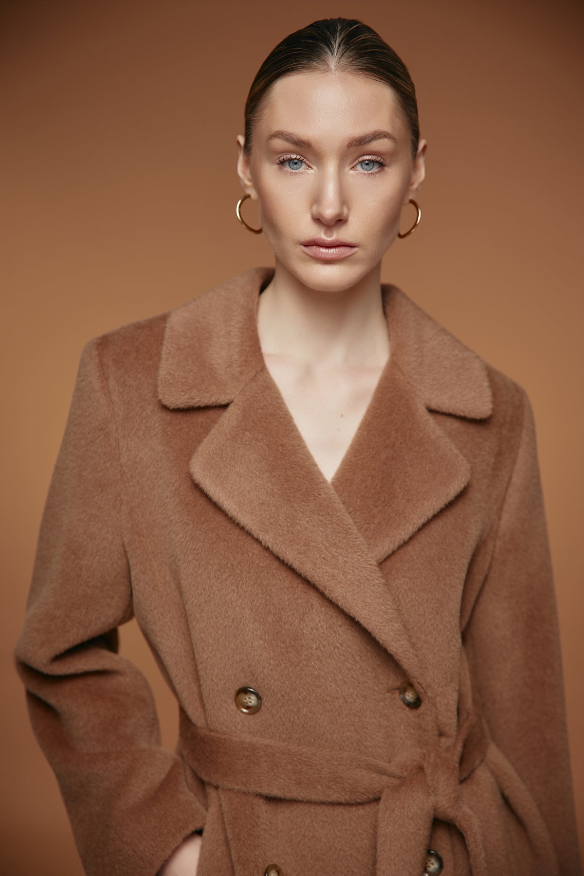 A woman wearing a camel brown double-breasted belted coat, styled with gold hoop earrings, against a solid warm brown studio background.