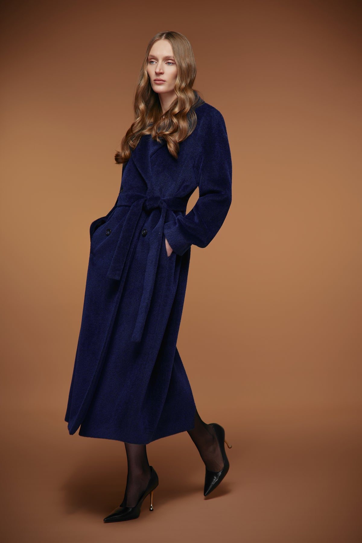A woman modeling a long navy blue velvet wrap coat with a tie belt, styled with black tights and black pointed-toe heels, against a solid warm brown studio background.