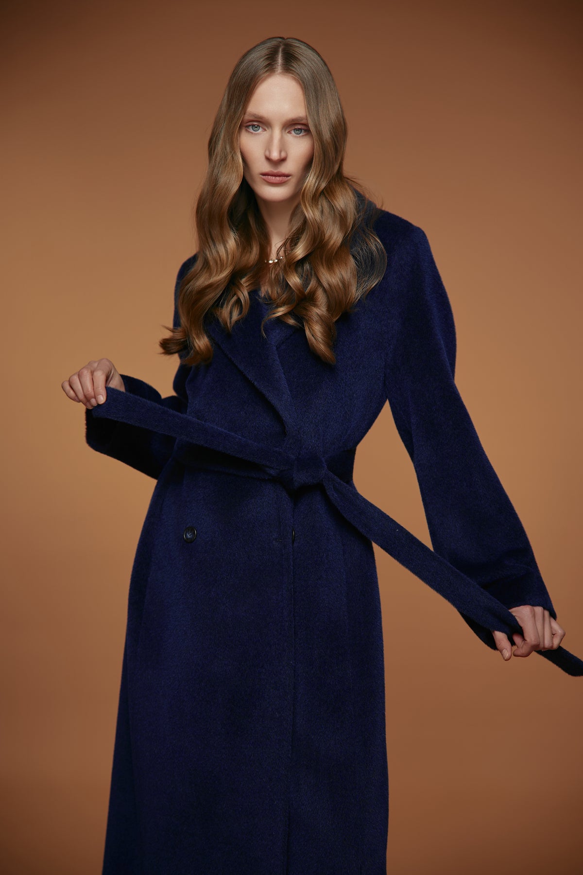A woman modeling a long navy blue coat with a matching waist tie, posed against a solid warm brown studio background.