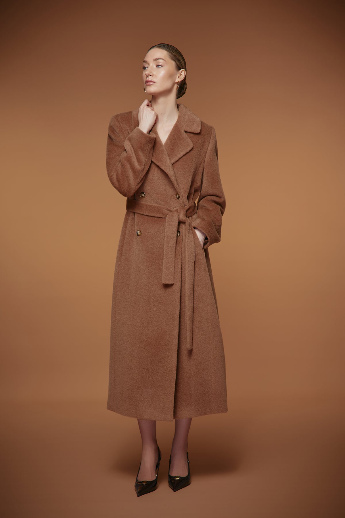 A woman models a long brown wool-blend coat with a matching waist tie belt, standing against a solid warm brown studio background.