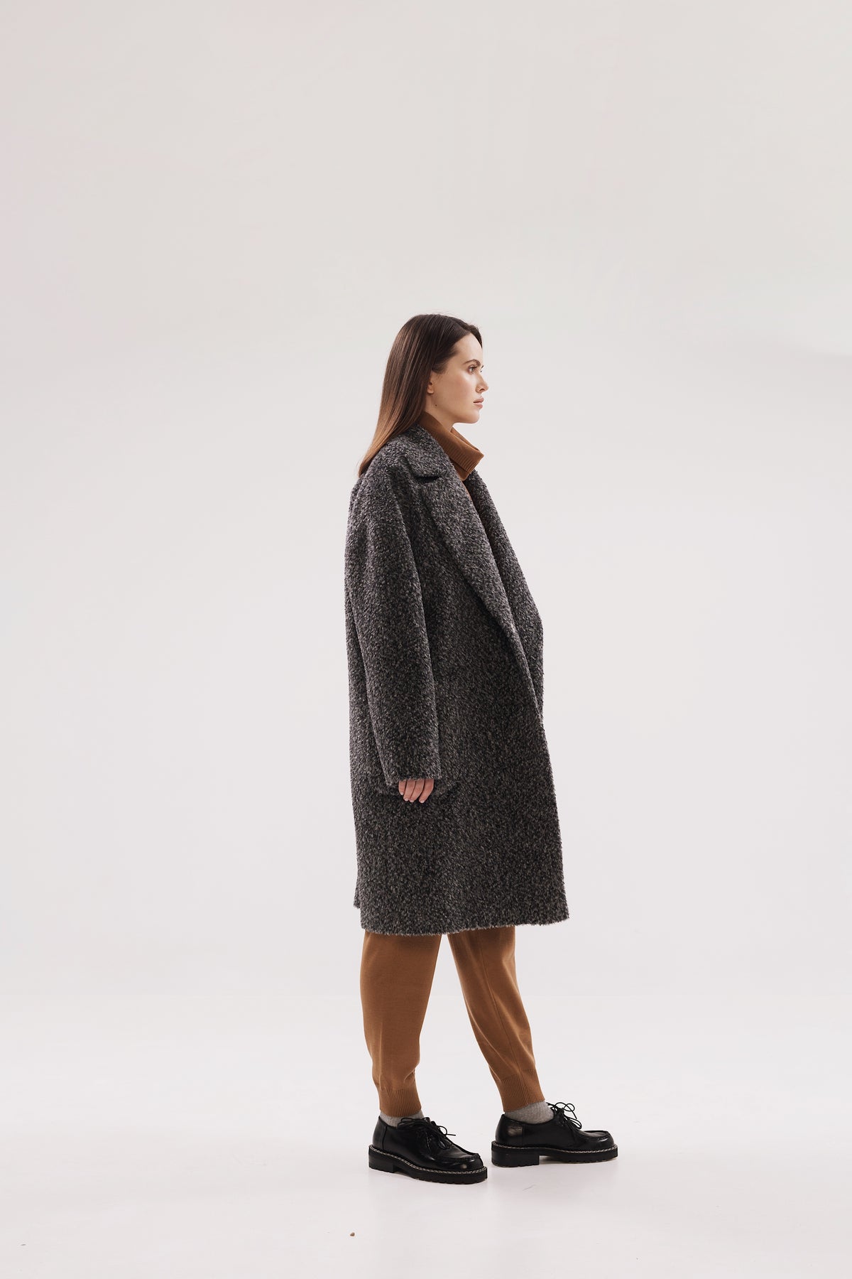 A woman stands in profile wearing a dark gray textured coat, brown pants, and black shoes against a plain light gray studio background.