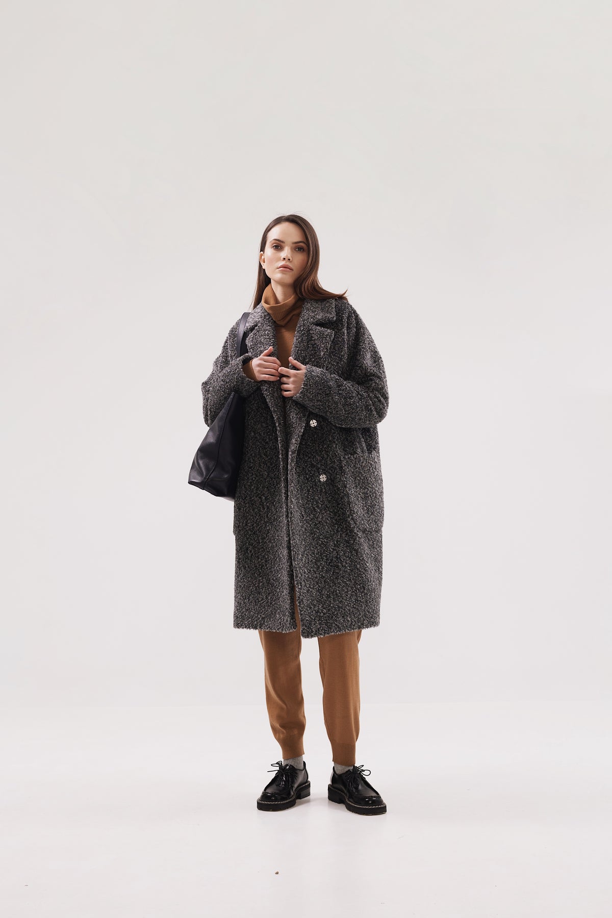 A woman modeling a long charcoal gray textured coat over a brown scarf and brown pants, standing against a plain light gray studio background.