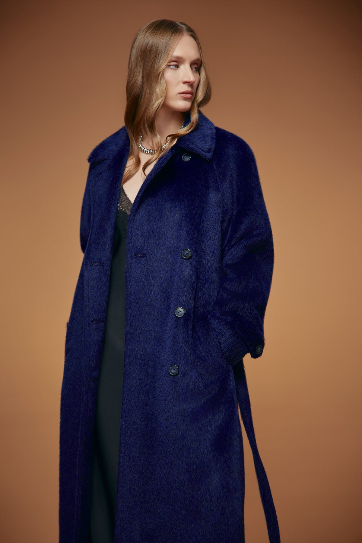 A woman modeling a long, dark blue textured coat over a black dress, set against a solid warm brown studio background.