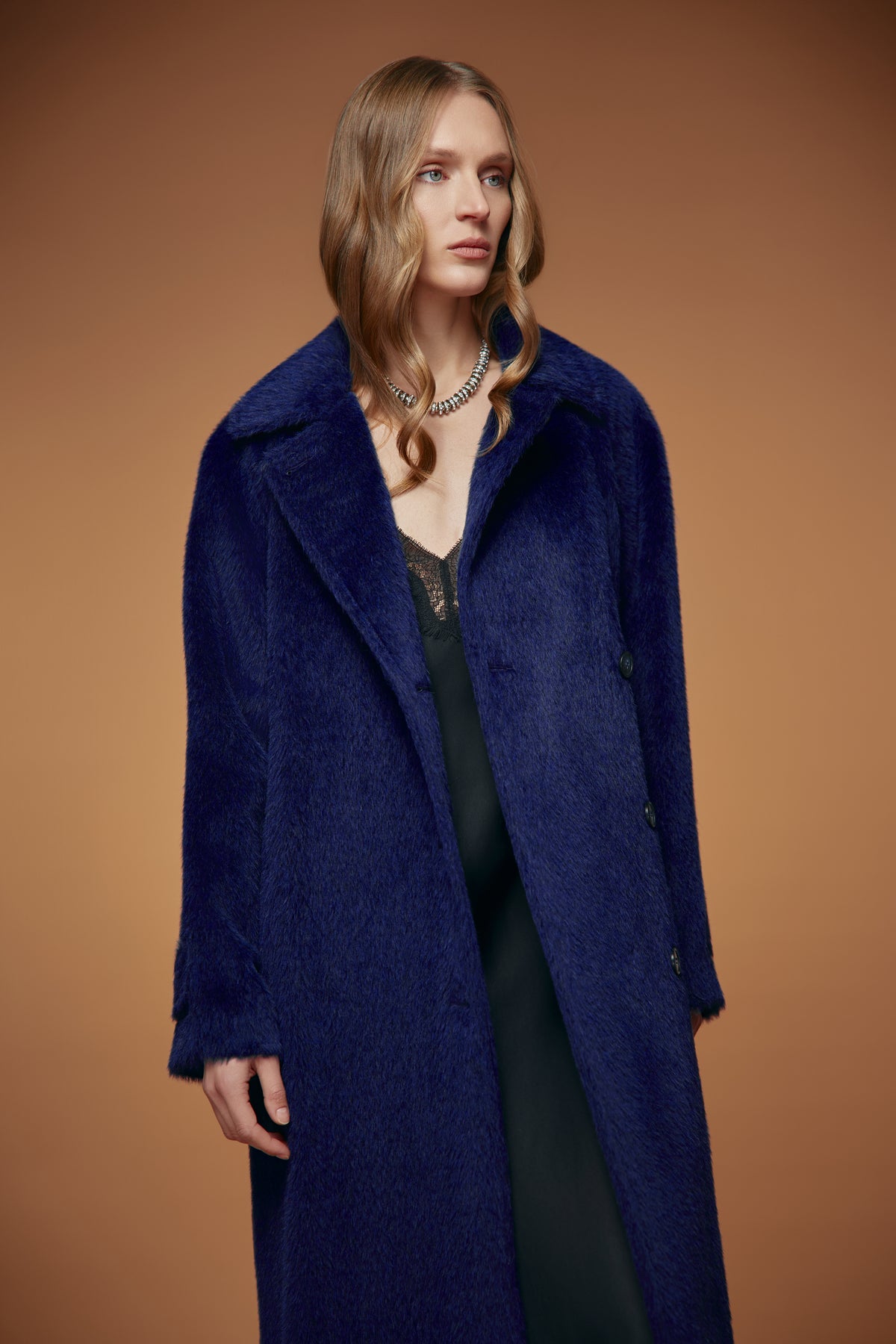 A woman modeling a long, plush navy blue coat over a black dress, set against a solid warm brown studio background.