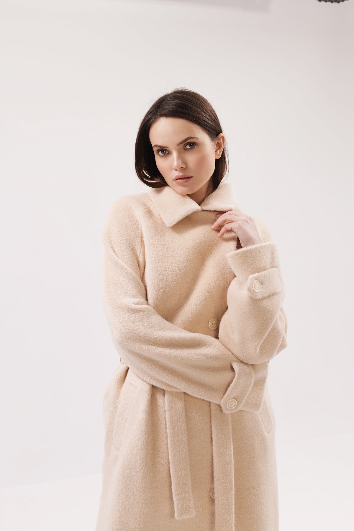 A woman wearing a long, soft beige wool coat with a collar and self-tie belt, posed against a clean white studio background.