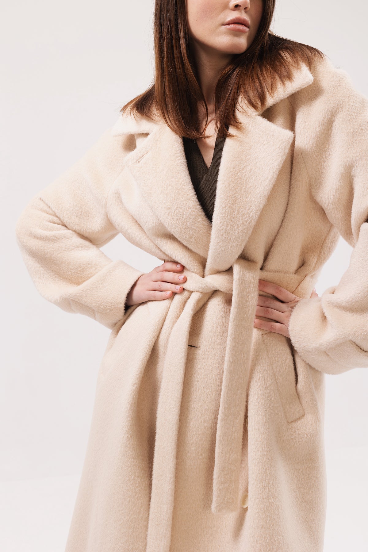 A woman wearing a plush, oversized beige coat with a tie belt, shown from the shoulders down against a plain light background.