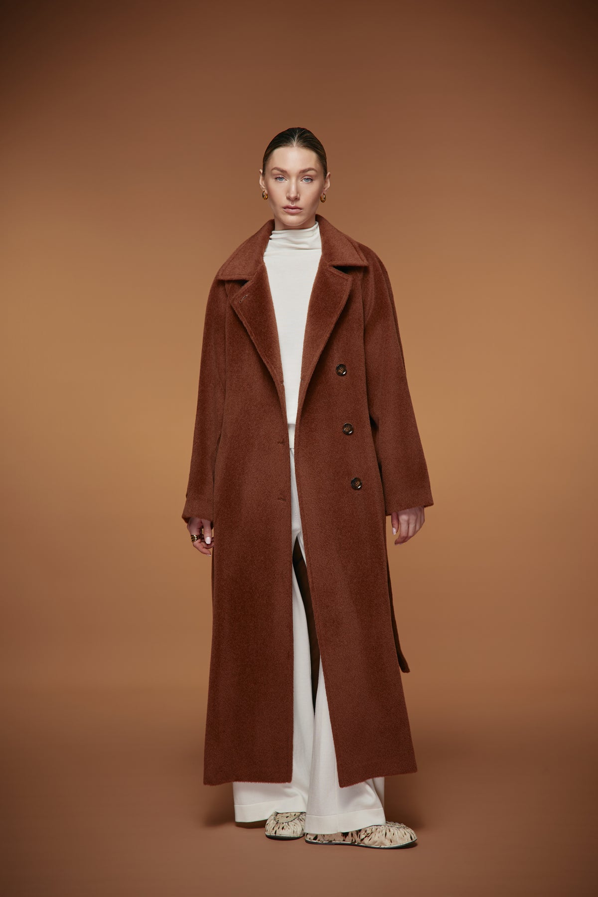 A model stands against a solid warm brown studio background wearing a long brown double-breasted coat over a white turtleneck and white wide-leg trousers.
