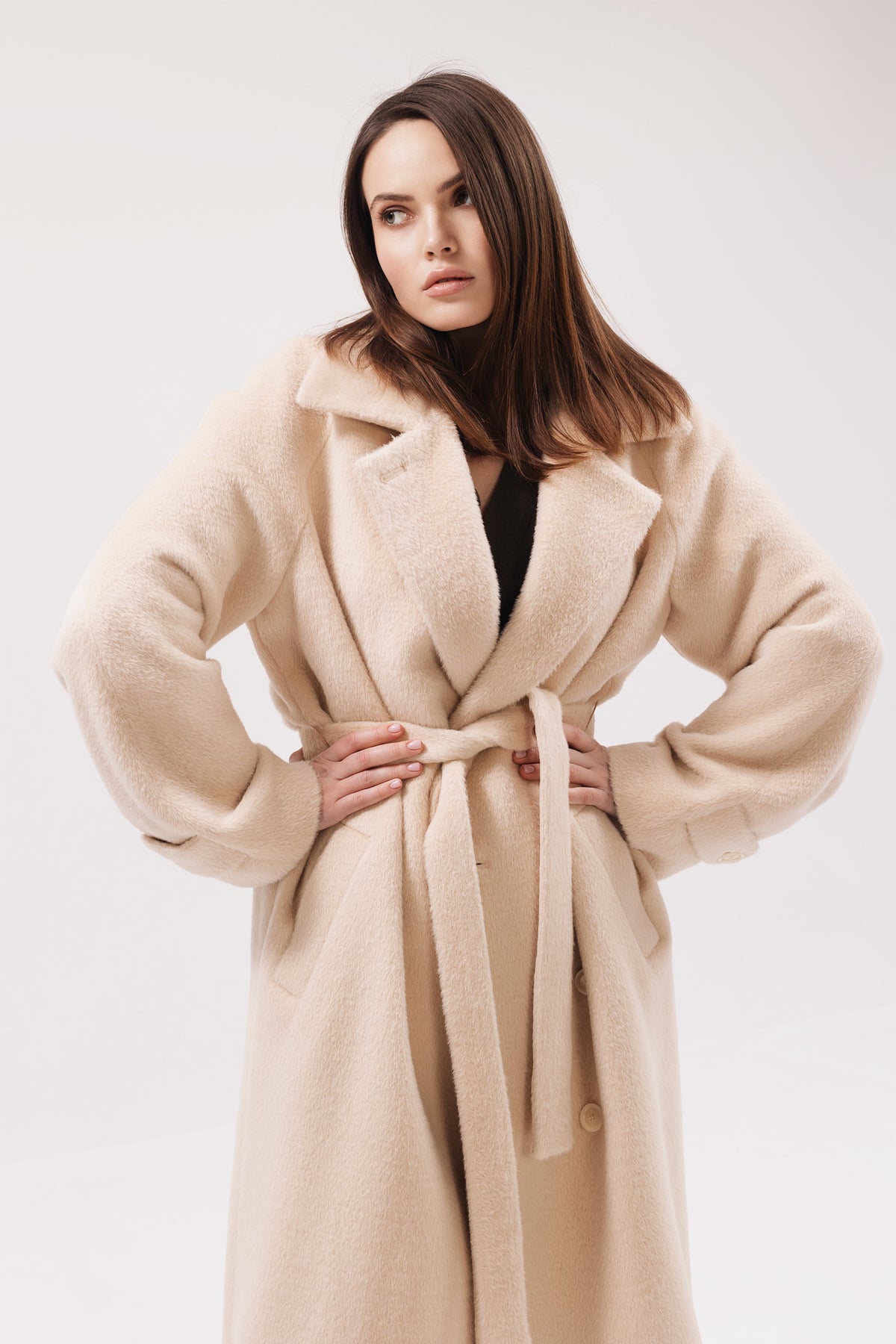 A woman modeling a long, plush beige coat with a tie belt, photographed against a clean white studio background.