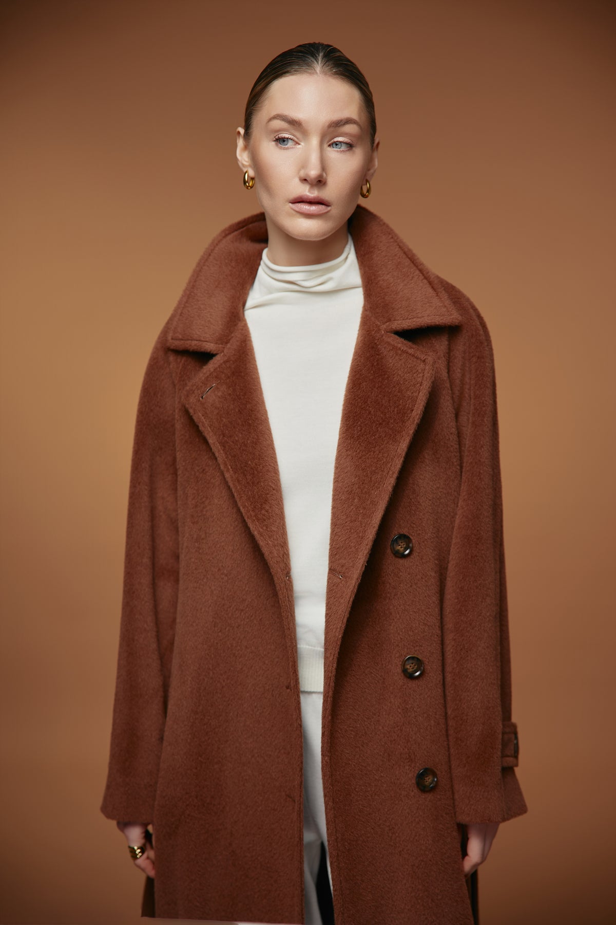 A woman wearing a brown double-breasted coat over a white turtleneck sweater, standing against a solid brown studio background.