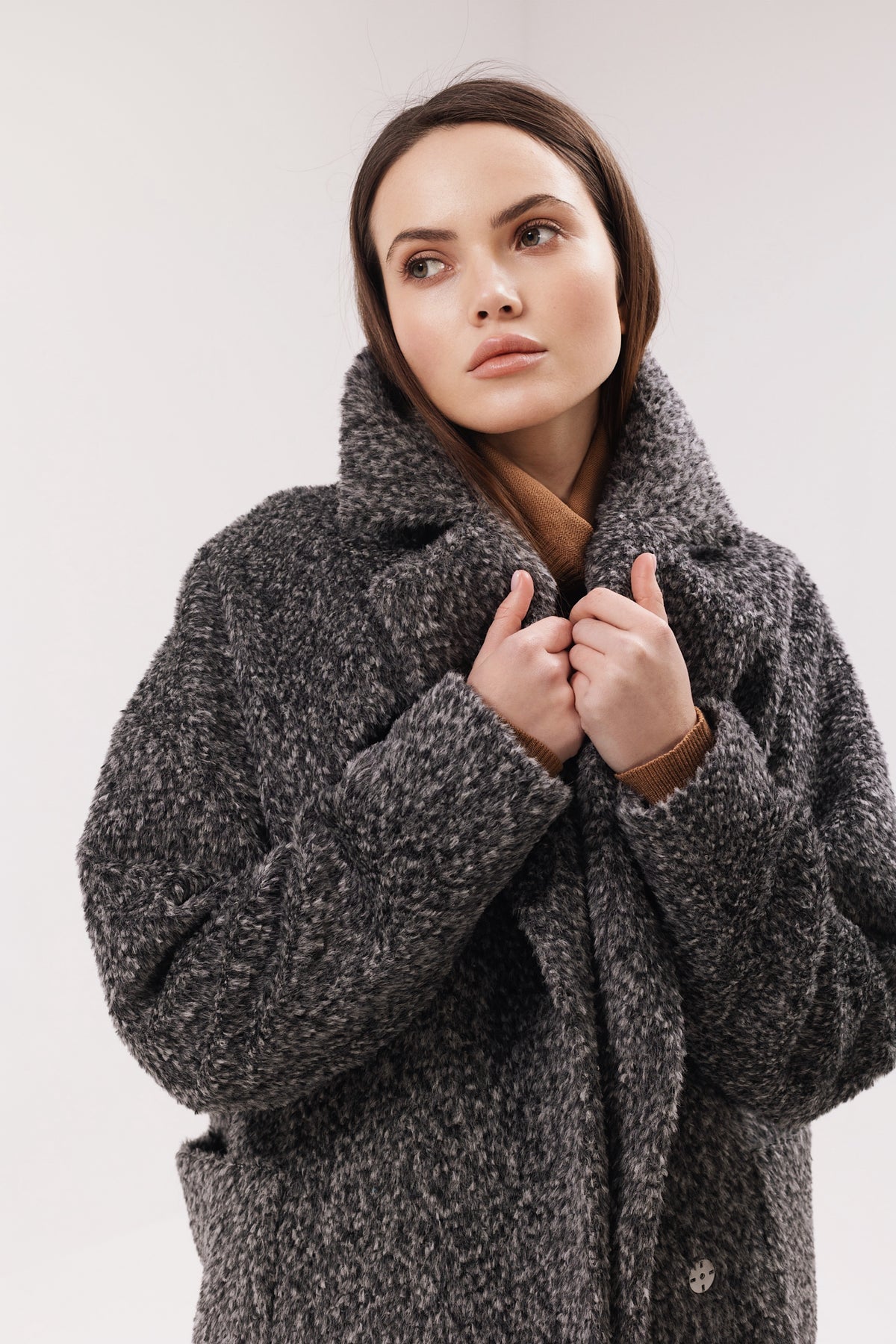 A woman wearing a chunky, textured dark gray bouclé coat with a high collar, posing against a light gray studio background.