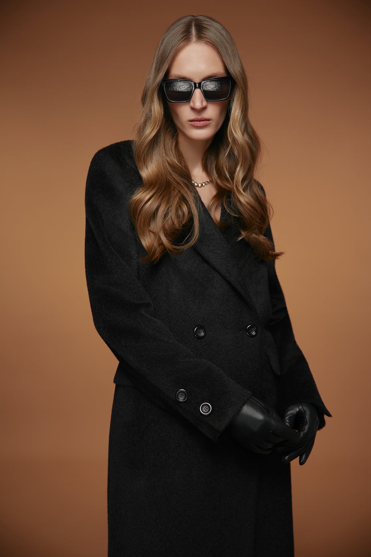 A woman wearing a long black double-breasted coat, black gloves, and black sunglasses, posed against a solid warm brown studio background.