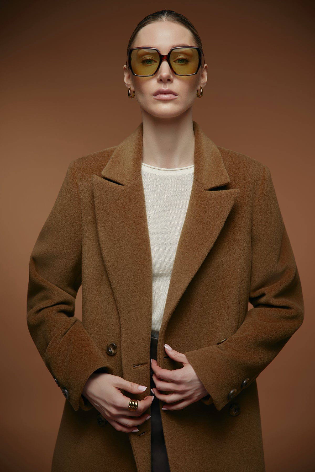 A model wearing a camel brown wool-blend coat over a cream top, styled with yellow-tinted sunglasses and gold jewelry, set against a solid warm brown studio background.