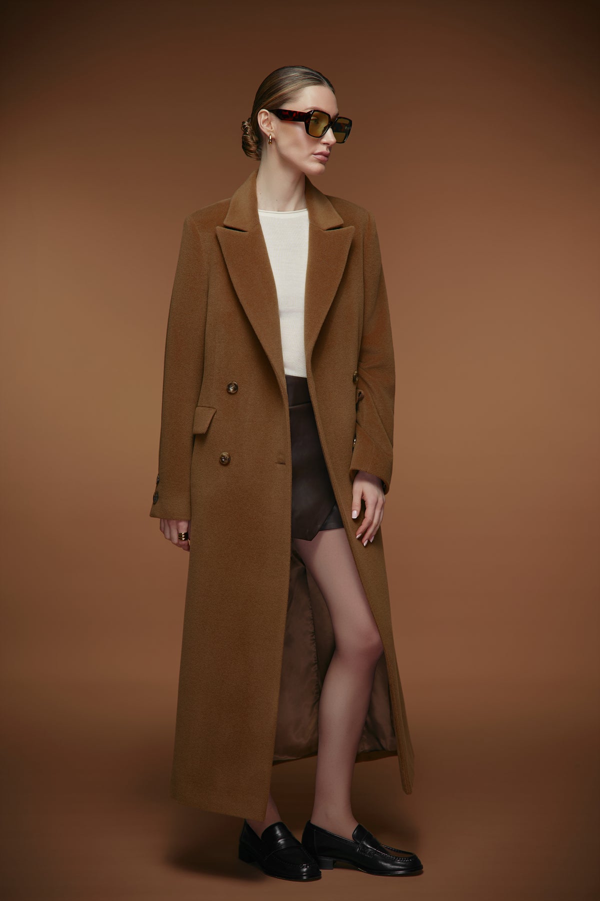 A model wearing a long brown double-breasted coat over a white top and black skirt, styled with black loafers and sunglasses, set against a solid brown studio background.