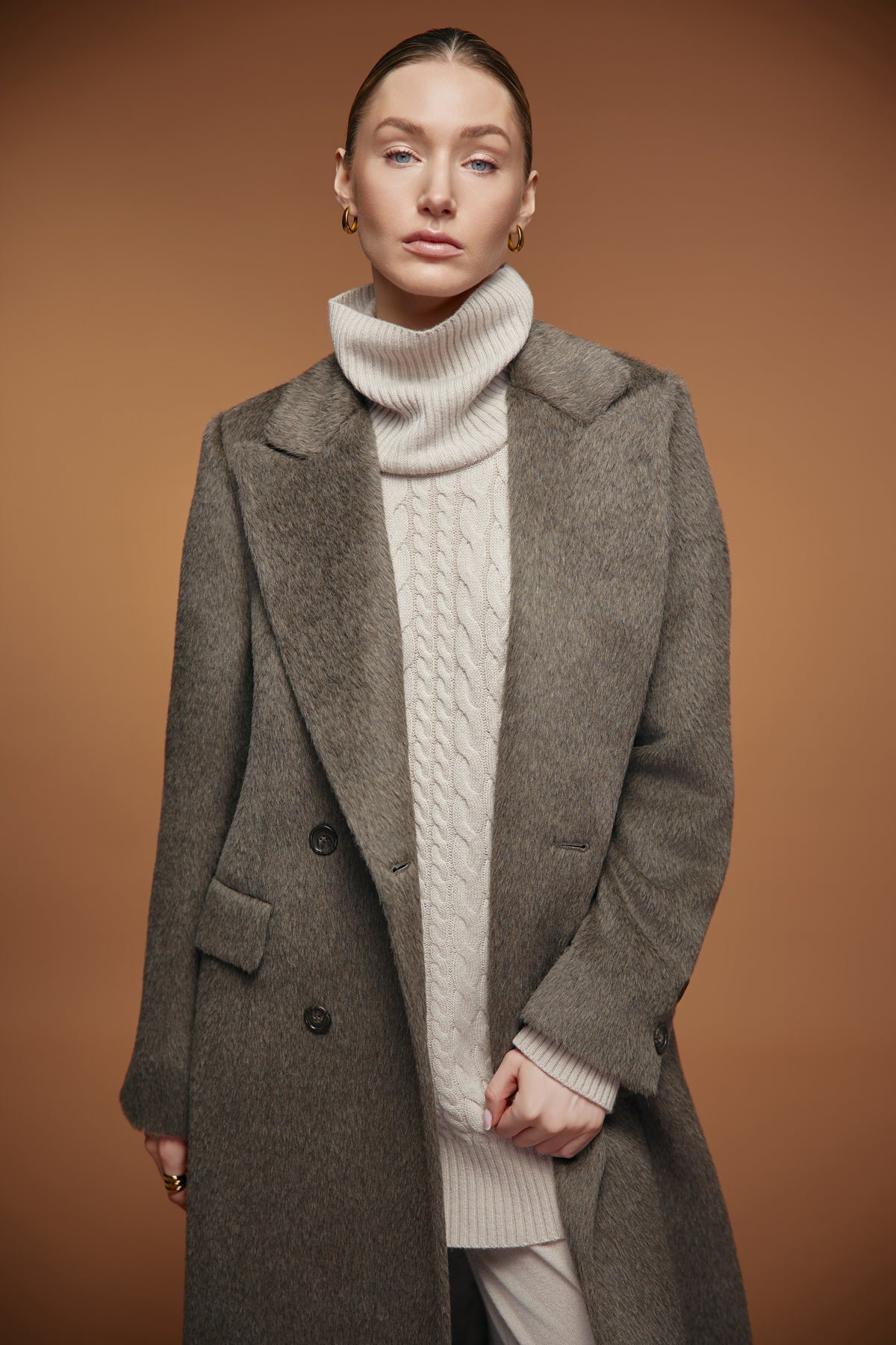 A woman modeling a long taupe-gray wool coat over a cream cable-knit turtleneck sweater, set against a solid warm brown studio background.