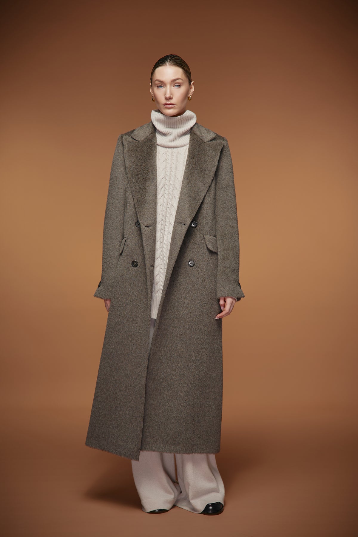 A model stands against a solid warm brown studio background wearing a long charcoal gray double-breasted overcoat over a cream cable-knit turtleneck sweater, light beige wide-leg trousers, and black pointed shoes.