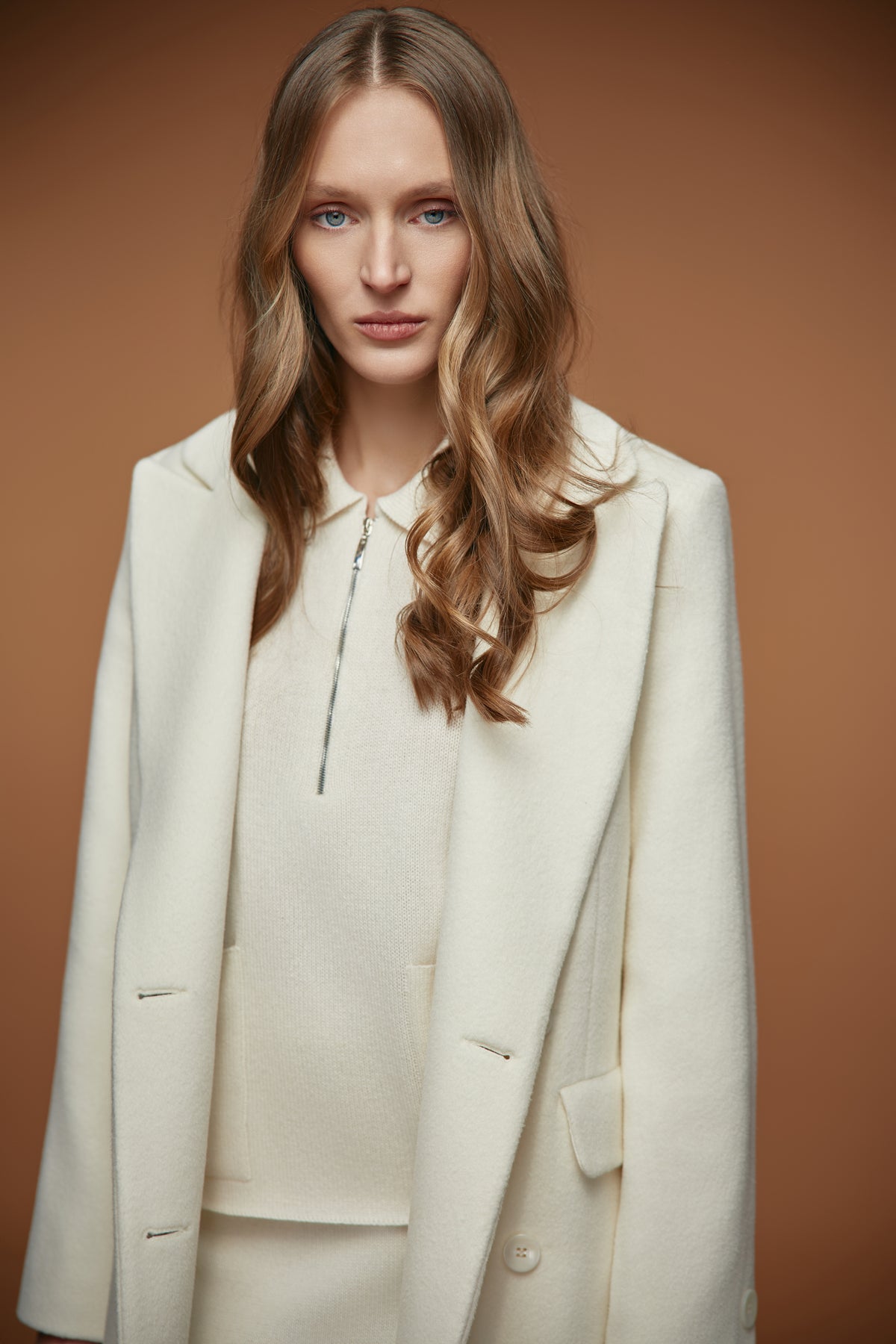 A woman with wavy light brown hair wearing a cream-colored tailored blazer over a matching cream zip-up top and trousers, posed against a solid warm brown studio background.