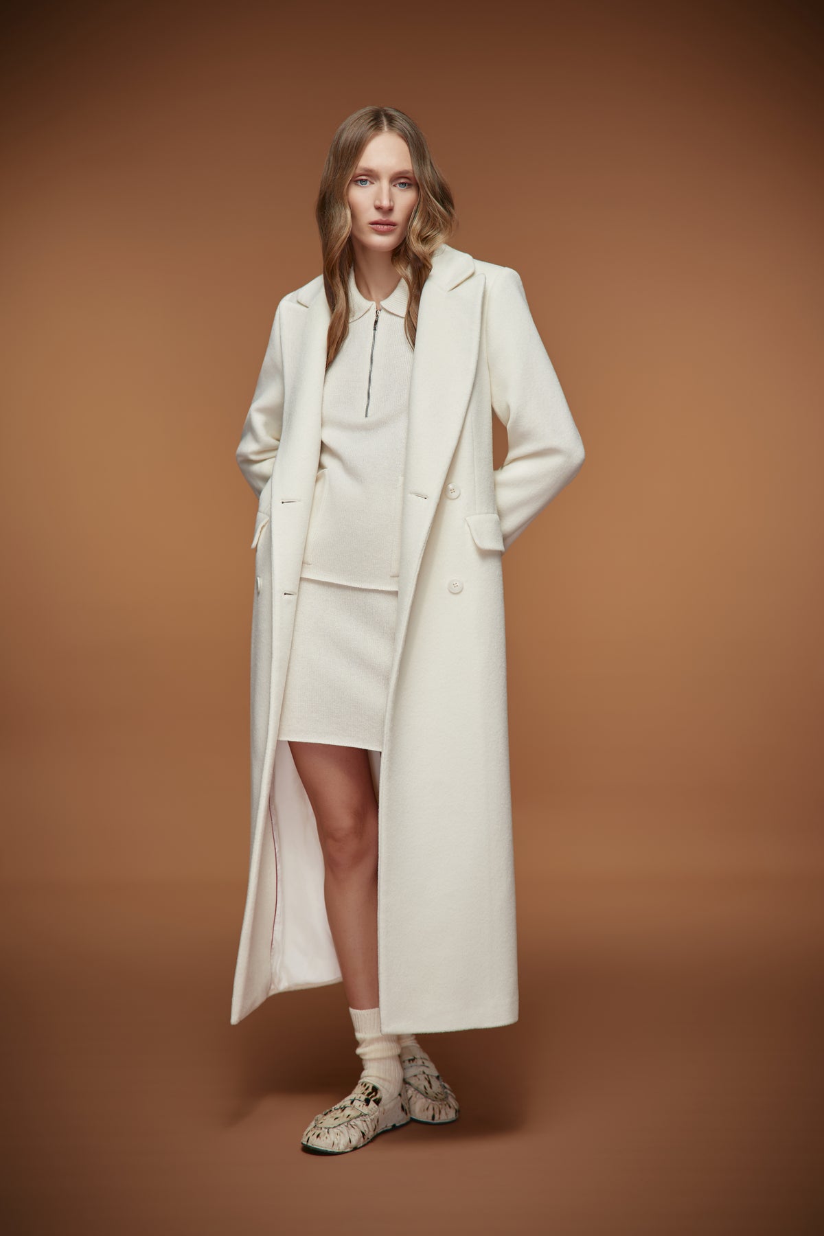 A model stands against a solid warm brown studio background wearing a long cream-colored wool-blend coat over a matching cream dress, paired with patterned knit socks and snakeskin sneakers.